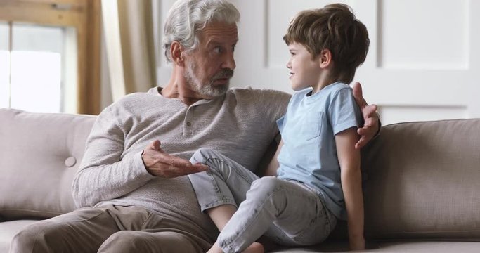 Joyful older senior grandfather telling fairy tale to cute little grandson, resting together in living room. Happy different male generations family enjoying funny talk chat conversation indoors.