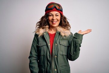 Middle age skier woman wearing snow sportswear and ski goggles over white background smiling cheerful presenting and pointing with palm of hand looking at the camera.