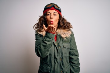 Middle age skier woman wearing snow sportswear and ski goggles over white background looking at the camera blowing a kiss with hand on air being lovely and sexy. Love expression.