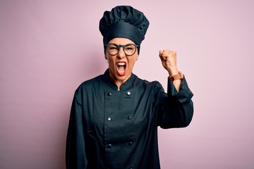 Middle age brunette chef woman wearing cooker uniform and hat over isolated pink background angry and mad raising fist frustrated and furious while shouting with anger. Rage and aggressive concept.