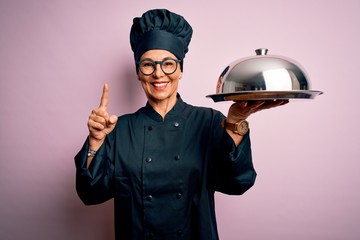 Middle age brunette chef woman wearing cooker uniform and hat holding tray dome surprised with an idea or question pointing finger with happy face, number one