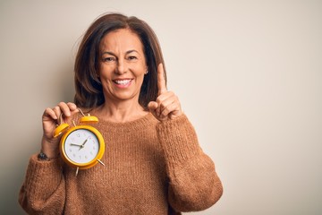 Middle age brunette woman holding clasic alarm clock over isolated background with a big smile on face, pointing with hand finger to the side looking at the camera.