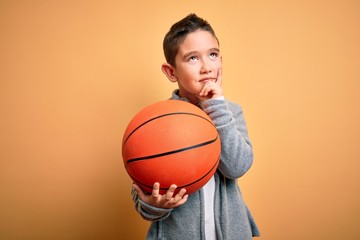 Young little boy kid playing with basketball game ball over isolated yellow background serious face thinking about question, very confused idea