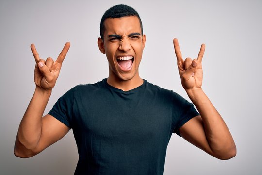 Young handsome african american man wearing casual t-shirt standing over white background shouting with crazy expression doing rock symbol with hands up. Music star. Heavy music concept.