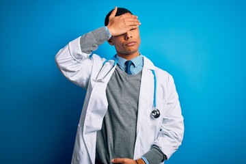 Handsome african american doctor man wearing coat and stethoscope over blue background covering eyes with hand, looking serious and sad. Sightless, hiding and rejection concept