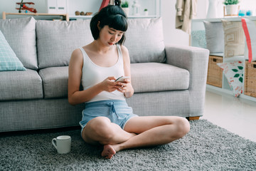 Portrait of smiling young asian korean cute woman sitting on floor at home with cellphone. attractive lady in shorts relaxing in morning on carpet text message sending online on mobile phone indoors