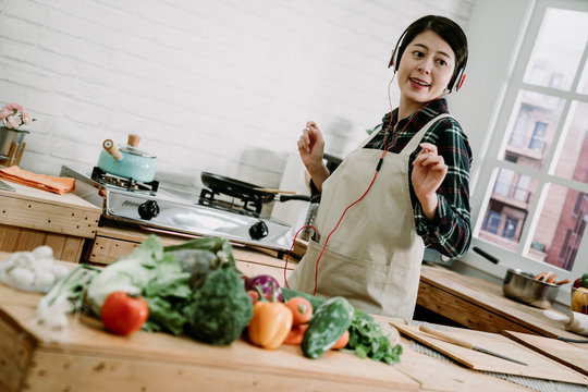 Enjoying her morning. Beautiful young asian japanese woman in headphones cooking salad and dancing while standing in kitchen at home. happy housewife in apron moving body with melody by earphones