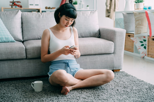 Portrait Of Smiling Young Asian Korean Cute Woman Sitting On Floor At Home With Cellphone. Attractive Lady In Shorts Relaxing In Morning On Carpet Text Message Sending Online On Mobile Phone Indoors