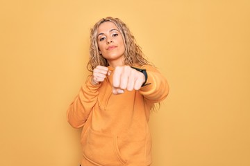 Young beautiful blonde sporty woman wearing casual sweatshirt over yellow background Punching fist to fight, aggressive and angry attack, threat and violence