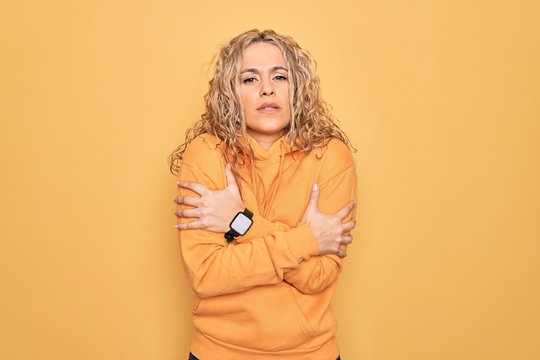 Young beautiful blonde sporty woman wearing casual sweatshirt over yellow background shaking and freezing for winter cold with sad and shock expression on face