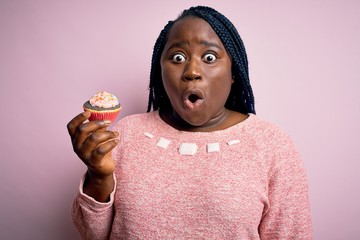 Young african american plus size woman with braids eating sweet chocolate cupcake scared in shock...