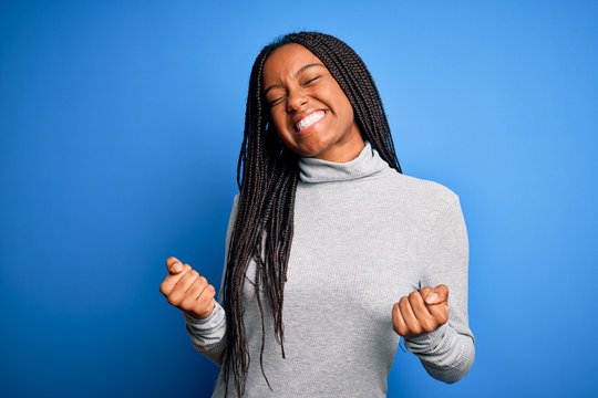 Young african american woman standing wearing casual turtleneck over blue isolated background very happy and excited doing winner gesture with arms raised, smiling and screaming for success. 