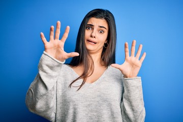 Young beautiful brunette woman wearing casual sweater standing over blue background afraid and terrified with fear expression stop gesture with hands, shouting in shock. Panic concept.