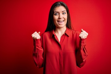 Young beautiful woman with blue eyes wearing casual shirt standing over red background celebrating surprised and amazed for success with arms raised and open eyes. Winner concept.