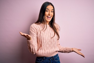 Young beautiful brunette woman wearing casual sweater over isolated pink background smiling cheerful with open arms as friendly welcome, positive and confident greetings
