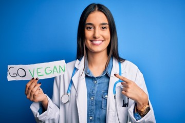Young beautiful brunette doctor woman wearing coat holding paper with go vegan message with surprise face pointing finger to himself