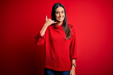 Young beautiful brunette woman wearing casual sweater over isolated red background smiling doing phone gesture with hand and fingers like talking on the telephone. Communicating concepts.