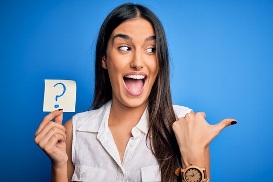 Young beautiful brunette woman holding paper with question mark symbol message pointing and showing with thumb up to the side with happy face smiling