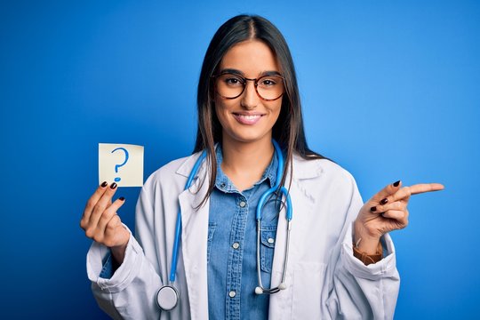 Young beautiful brunette doctor woman holding paper with question mark symbol message very happy pointing with hand and finger to the side