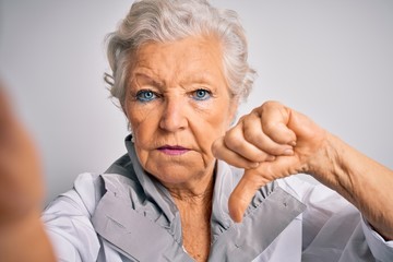 Senior beautiful grey-haired woman making selfie by camera over isolated white background with angry face, negative sign showing dislike with thumbs down, rejection concept