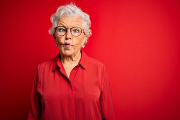 Senior beautiful grey-haired woman wearing casual shirt and glasses over red background making fish...