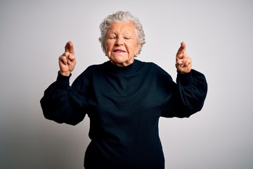 Senior beautiful woman wearing casual black sweater standing over isolated white background gesturing finger crossed smiling with hope and eyes closed. Luck and superstitious concept.