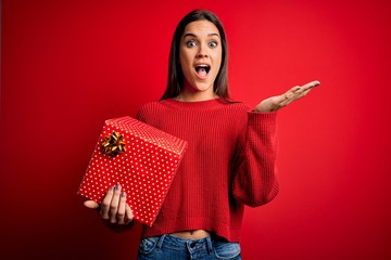 Young beautiful brunette woman holding birthday gift over isolated red background very happy and excited, winner expression celebrating victory screaming with big smile and raised hands