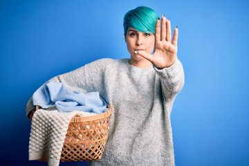 Young woman with blue fashion hair doing domestic chores holding wicker laundry basket with open hand doing stop sign with serious and confident expression, defense gesture