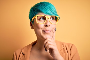 Young beautiful woman with blue fashion hair wearing casual glasses over yellow background serious...