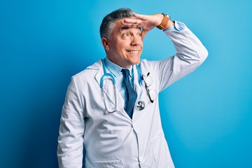 Middle age handsome grey-haired doctor man wearing coat and blue stethoscope very happy and smiling looking far away with hand over head. Searching concept.