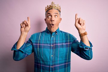 Middle age handsome grey-haired man wearing golden crown of king over pink background amazed and...
