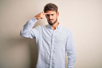 Young handsome man with beard wearing striped shirt standing over white background pointing unhappy to pimple on forehead, ugly infection of blackhead. Acne and skin problem