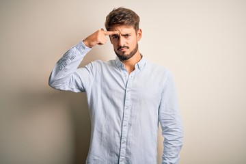 Young handsome man with beard wearing striped shirt standing over white background pointing unhappy...