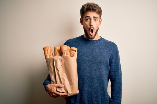 Young man holding paper bag with bread standing over isolated red bakground scared in shock with a surprise face, afraid and excited with fear expression
