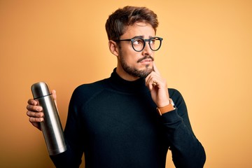 Young man on vacation wearing glasses holding metallic thermo wih water serious face thinking about...