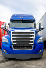 Bright blue big rig semi truck with chrome grille standing in row with another semi trucks with semi trailers on truck stop parking lot for rest