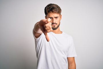 Young handsome man with beard wearing casual t-shirt standing over white background looking unhappy and angry showing rejection and negative with thumbs down gesture. Bad expression.