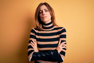 Young beautiful brunette woman wearing striped turtleneck sweater over yellow background skeptic and nervous, disapproving expression on face with crossed arms. Negative person.