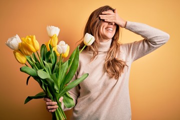Young beautiful brunette woman holding bouquet of yellow tulips over isolated background smiling and laughing with hand on face covering eyes for surprise. Blind concept.