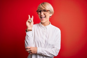 Young blonde business woman with short hair wearing glasses over red background smiling with happy face winking at the camera doing victory sign. Number two.