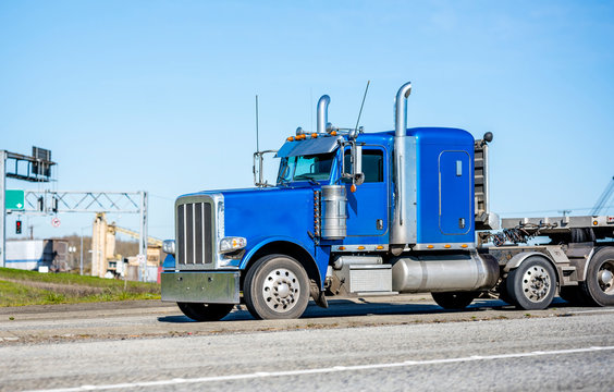 Classic blue big rig semi truck with tall exhaust pipes transporting cargo on flat bed semi trailer running on the road in industrial area