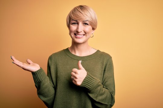 Young blonde woman with modern short hair wearing casual sweater over yellow background Showing palm hand and doing ok gesture with thumbs up, smiling happy and cheerful