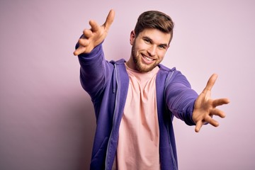 Naklejka premium Young blond man with beard and blue eyes wearing purple sweatshirt over pink background looking at the camera smiling with open arms for hug. Cheerful expression embracing happiness.