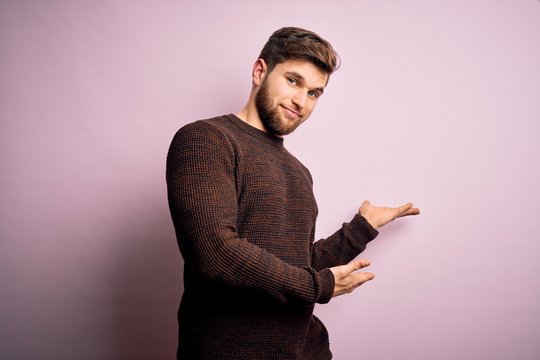 Young blond man with beard and blue eyes wearing casual sweater over pink background Inviting to enter smiling natural with open hand