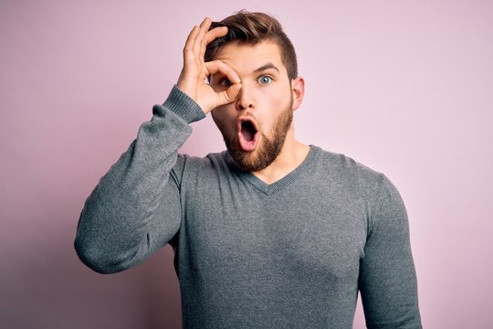 Young handsome blond man with beard and blue eyes wearing casual sweater doing ok gesture shocked with surprised face, eye looking through fingers. Unbelieving expression.