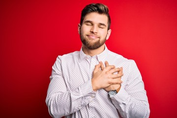 Young blond businessman with beard and blue eyes wearing elegant shirt over red background smiling with hands on chest with closed eyes and grateful gesture on face. Health concept.