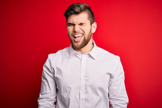 Young blond businessman with beard and blue eyes wearing elegant shirt over red background winking looking at the camera with sexy expression, cheerful and happy face.
