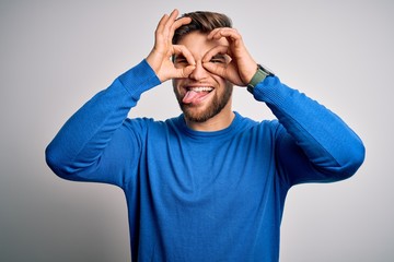 Young handsome blond man with beard and blue eyes wearing casual sweater doing ok gesture like binoculars sticking tongue out, eyes looking through fingers. Crazy expression.