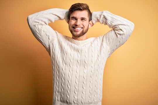 Young blond man with beard and blue eyes wearing white sweater over yellow background relaxing and stretching, arms and hands behind head and neck smiling happy