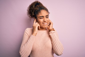 Young beautiful african american girl wearing casual sweater standing over pink background covering ears with fingers with annoyed expression for the noise of loud music. Deaf concept.